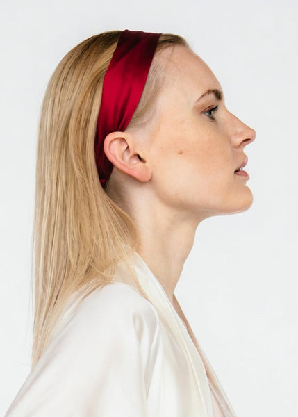 A blonde woman in profile wearing a wide, deep red silk headband and a white satin robe, set against a plain light gray background.