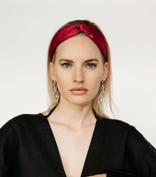 A woman wearing a deep red silk knotted headband and large silver hoop earrings, dressed in a black top, photographed against a plain light gray background.