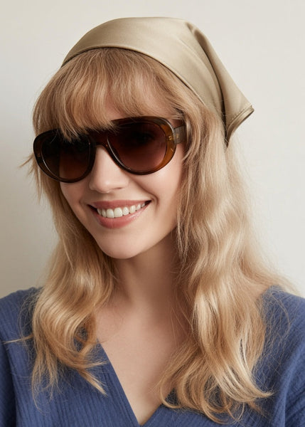 A smiling blonde woman wearing large dark sunglasses, a beige silk headscarf, and a blue textured top against a plain light background.