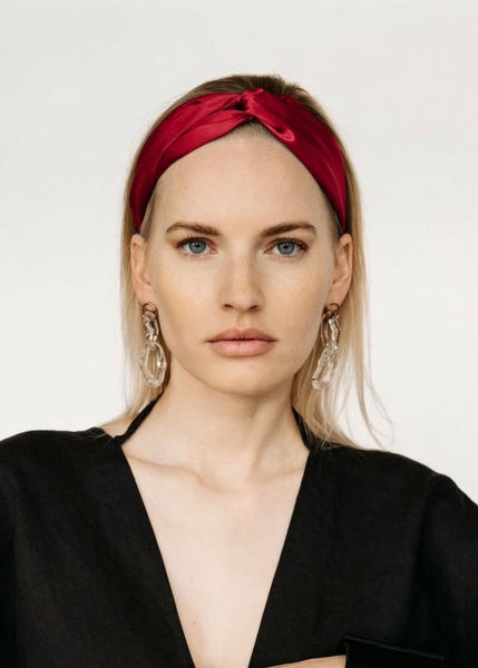 A woman wearing a deep redsilkknotted headband and large silver hoop earrings, dressed in a black top, photographed against a plain light gray background.